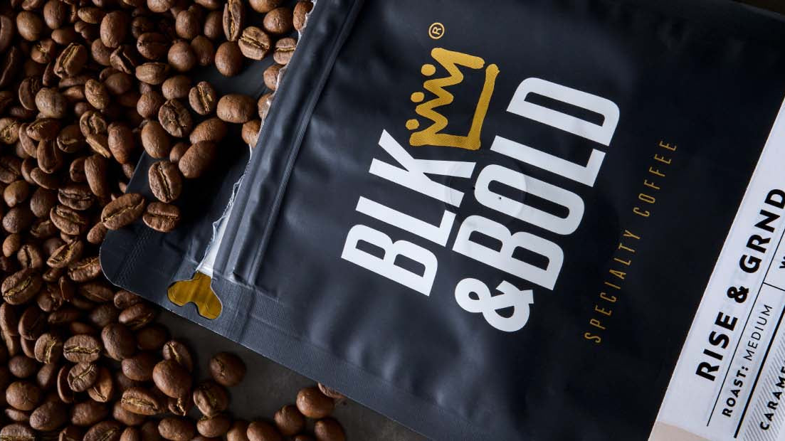 A black stand-up pouch of Blk & Bold Specialty Coffee, labeled 'Rise & GRND', shown diagonally over a dense pile of roasted coffee beans.